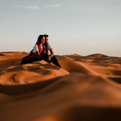 Couple Sitting On Top Of A Dune In The Sahara Desert Watching The Sunset stock photo